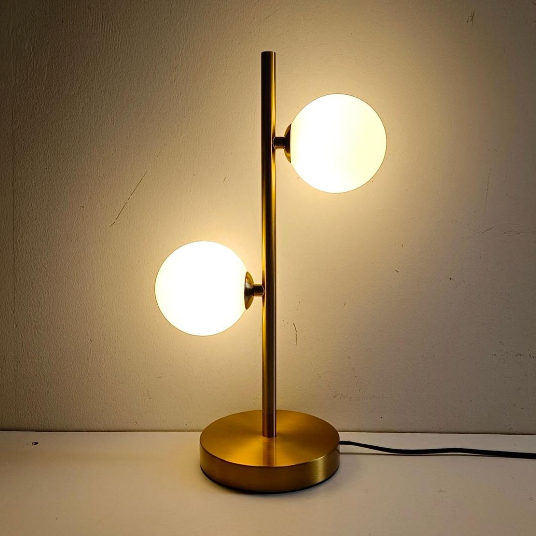 Abajur de mesa dourado com 2 globos de vidro branco acesos contra parede clara - Vittara Decor