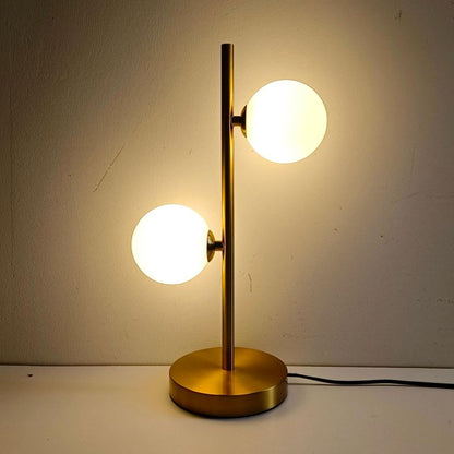 Abajur de mesa dourado com 2 globos de vidro branco acesos contra parede clara - Vittara Decor