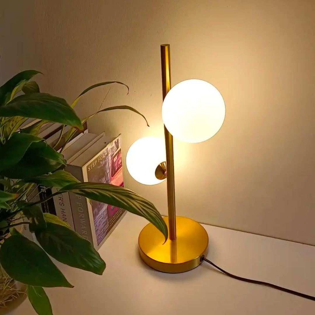 Abajur de mesa dourado com globos de vidro aceso ao lado de planta e livros - Vittara Decor