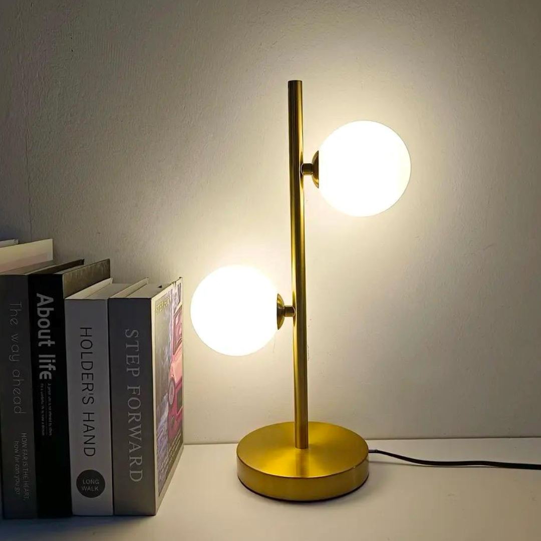 Abajur de mesa dourado de 2 globos de vidro aceso ao lado de livros na cabeceira - Vittara Decor