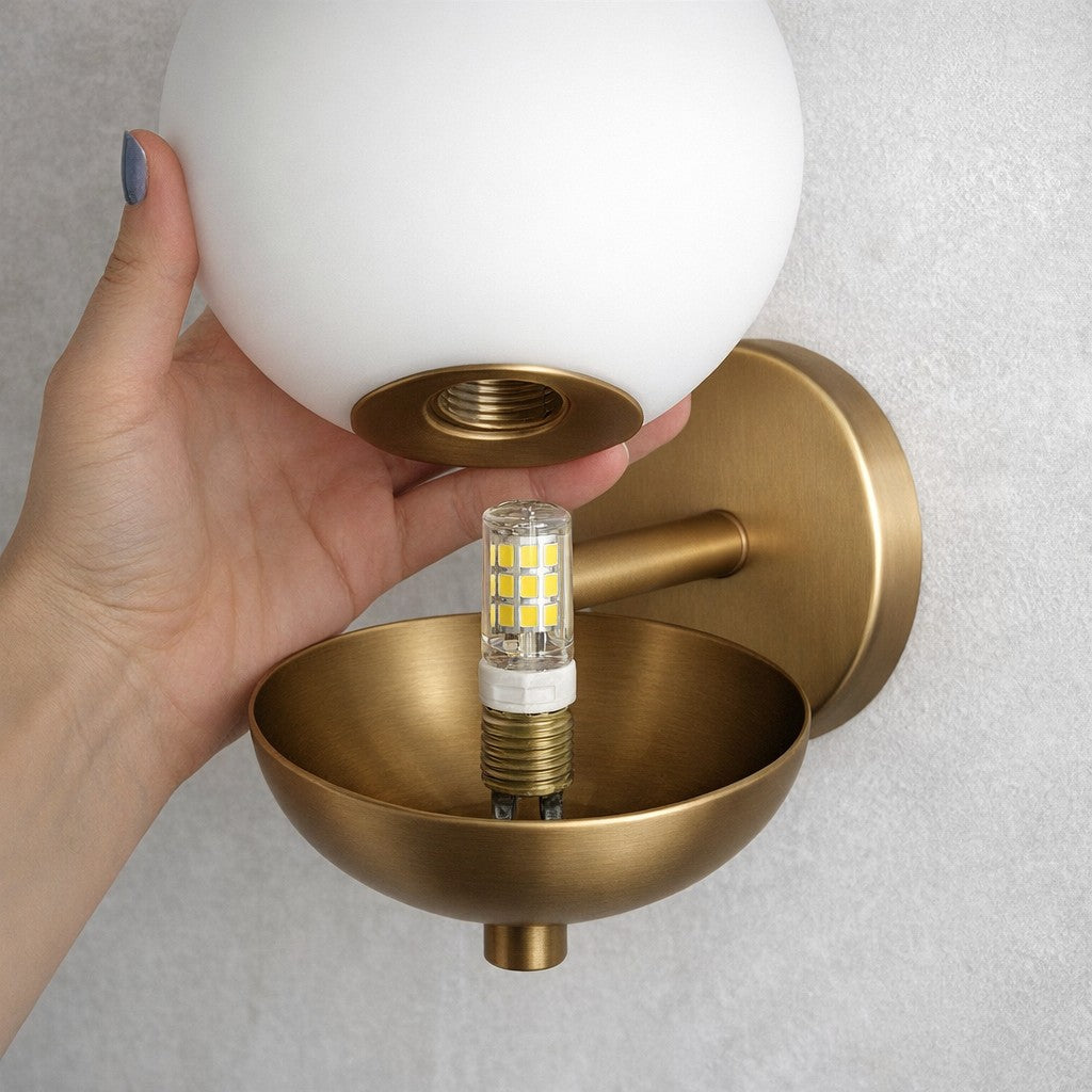 Mão retirando o globo opalino da arandela e mostrando a lâmpada G9 de pino - Vittara Decor