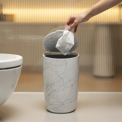 Lixeira multiuso mármore branco com tampa aberta e mão descartando papel em banheiro com painel dourado - Vittara Decor