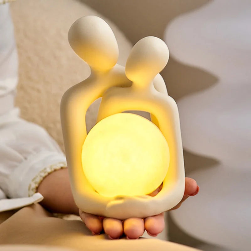 Luminária de casal abraçado nas mãos com esfera de luz LED acesa - Vittara Decor